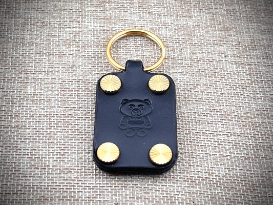Black leather key fob with gold studs and a bear emblem on a textured gray background
