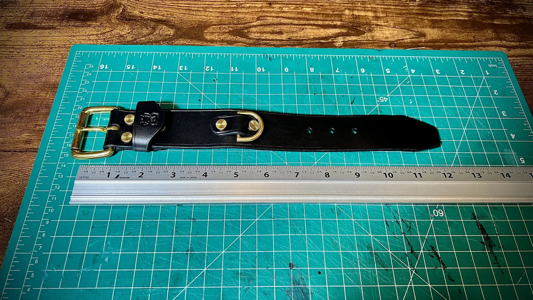 Top down view of a handcrafted black leather cuff with solid brass hardware laid flat on a green cutting mat alongside a metal ruler.