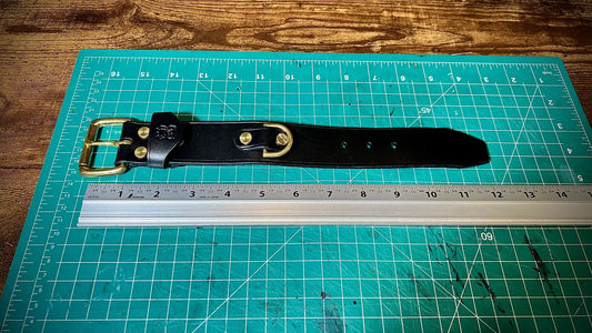 Top down view of a handcrafted black leather cuff with solid brass hardware laid flat on a green cutting mat alongside a metal ruler.