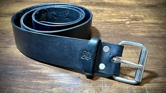 Studio view of a black vegetable-tanned leather belt with solid nickel steel roller buckle and dual rivets on a wood workspace.