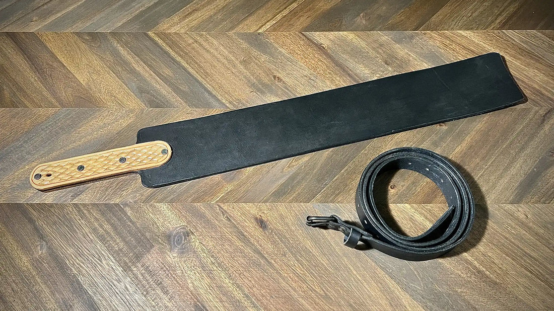 Studio view of a wide black leather paddle implement featuring a textured natural leather handle overlay alongside a matching rolled belt.