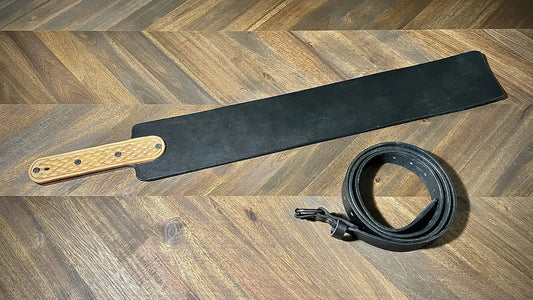 Studio view of a wide black leather paddle implement featuring a textured natural leather handle overlay alongside a matching rolled belt.