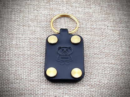 Black leather key fob with gold studs and a bear emblem on a textured gray background