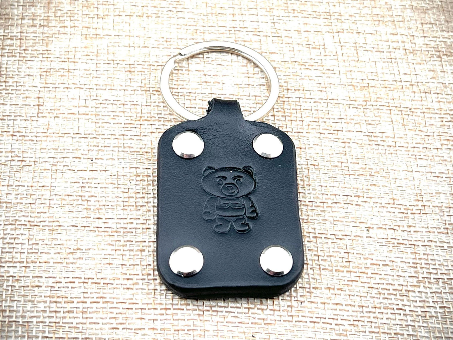 Black leather keychain with bear emblem and silver rivets on a textured beige background