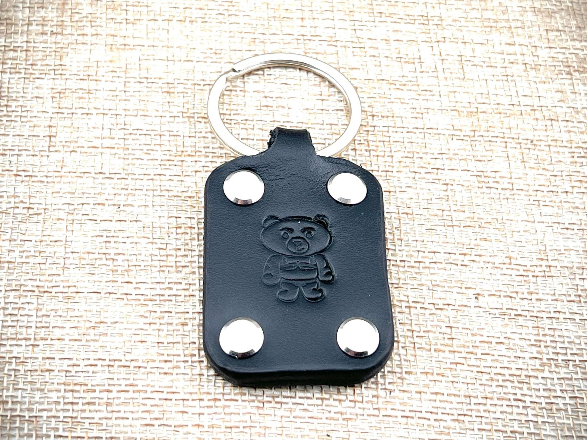 Black leather keychain with bear emblem and silver rivets on a textured beige background
