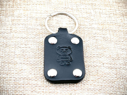 Black leather keychain with bear emblem and silver rivets on a textured beige background