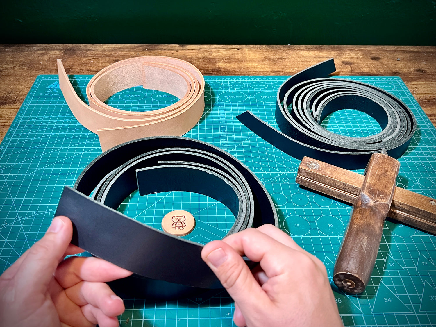 Hands holding a black leather strip on a cutting mat with leather strips and tools in the background.