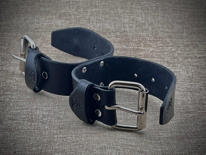 Two black leather bondage cuffs s with silver buckles on a textured gray background