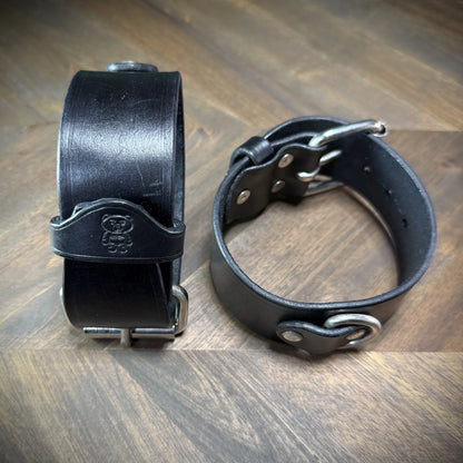 Front view of buckled 1.75" black leather cuffs standing on edge, highlighting an embossed bear brand mark and internal steel rivet assembly.
