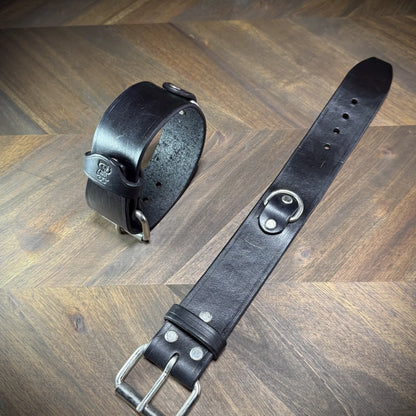 Overhead view of a pair of Knotty Oso 1.75" black leather cuffs with steel buckles and centered steel D-rings for consensual positioning.