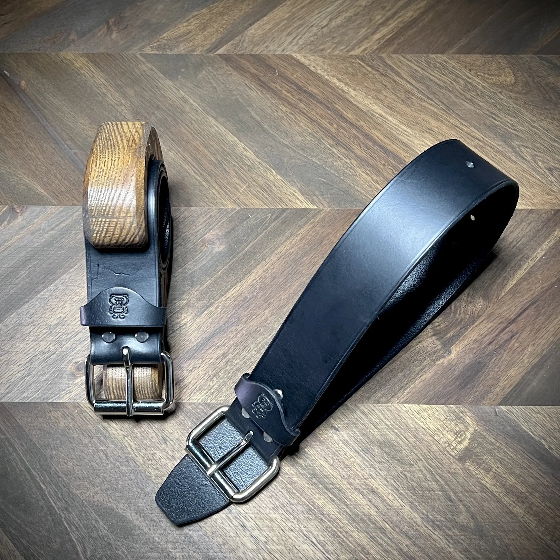 Diagonal view of a pair of Knotty Oso restraints bench-crafted from 8oz vegetable-tanned leather, showcasing the smooth strap and steel dual-prong buckles.