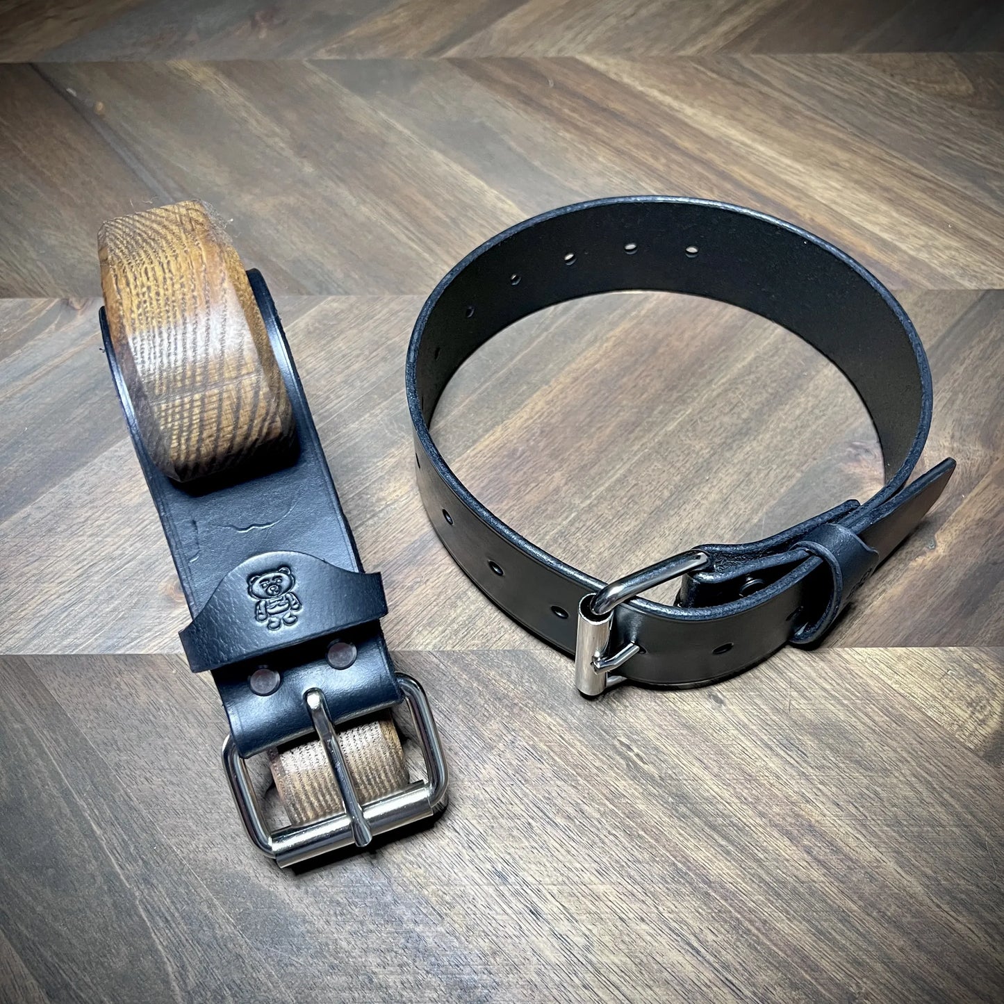 Overhead view of a pair of Knotty Oso restraints handcrafted from black leather, highlighting one rolled belt and one fastened with a steel buckle.