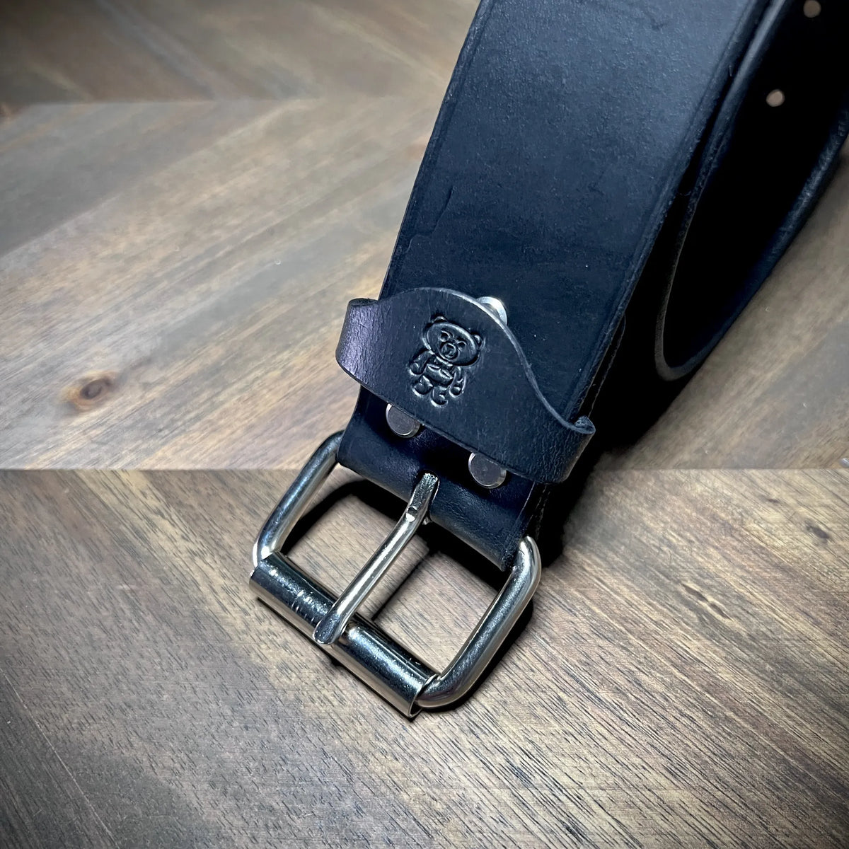 Macro view of a handcrafted black leather belt end showcasing a polished steel dual-prong buckle, dual rivets, and a debossed bear logo loop.
