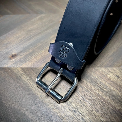 Macro view of a handcrafted black leather belt end showcasing a polished steel dual-prong buckle, dual rivets, and a debossed bear logo loop.