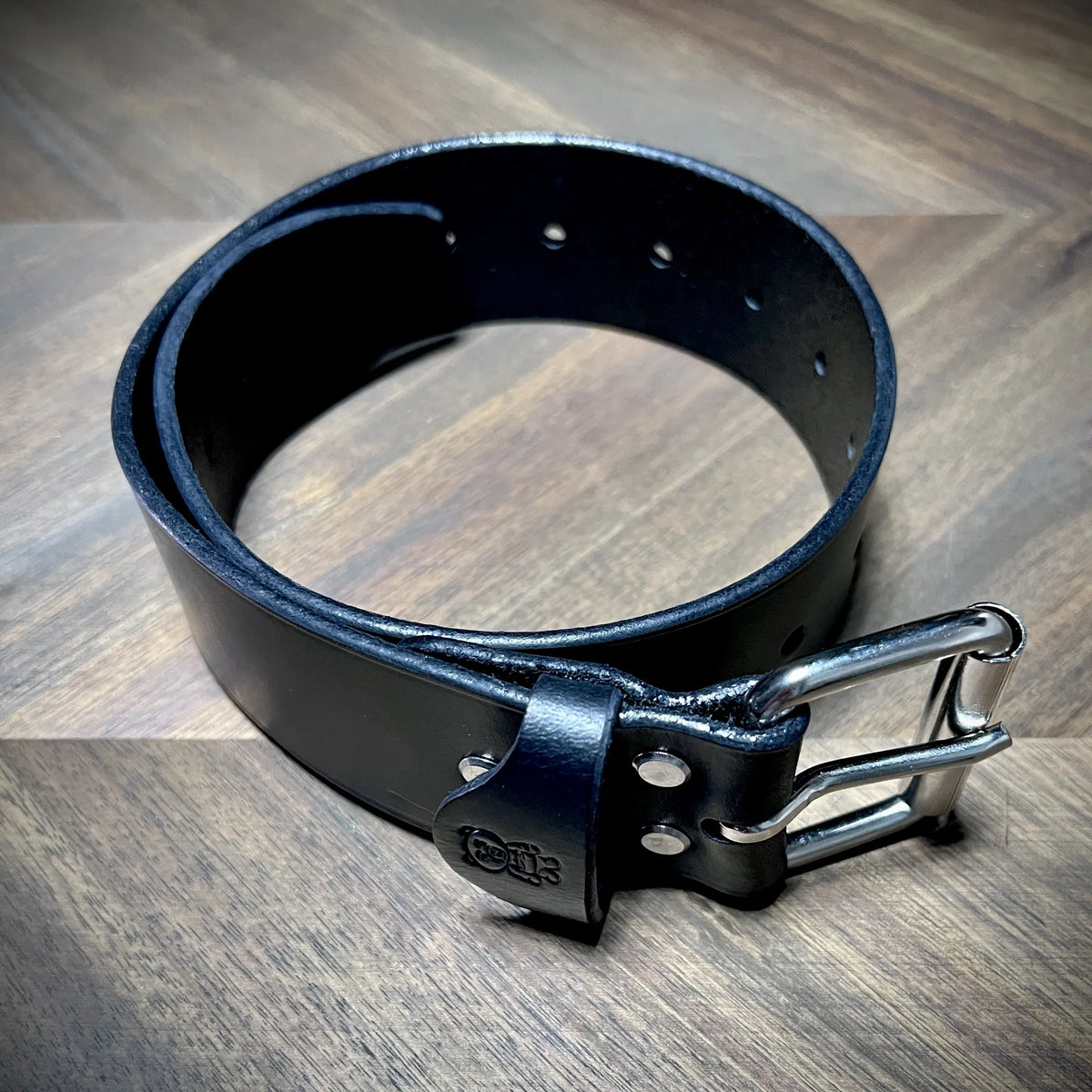 High-angle view of a 1.75" black leather restraint coiled free-standing, showcasing a dual-prong steel buckle and parallel adjustment hole rows.