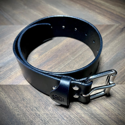 High-angle view of a 1.75" black leather restraint coiled free-standing, showcasing a dual-prong steel buckle and parallel adjustment hole rows.