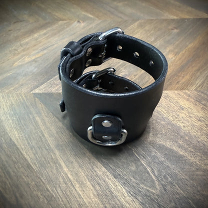 Hero tilt view of a closed 3.5" black leather bondage cuff, emphasizing its thick structural curve and centered steel D-ring assembly.