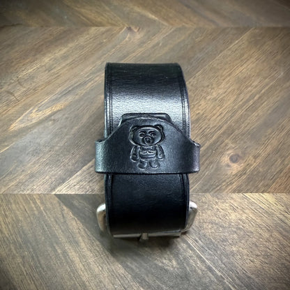 Vertical front view of a buckled 1.5" black leather cuff focusing on a centered debossed cartoon bear logo over the smooth leather strap.