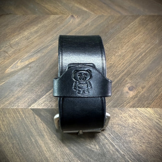 Vertical front view of a buckled 1.5" black leather cuff focusing on a centered debossed cartoon bear logo over the smooth leather strap.