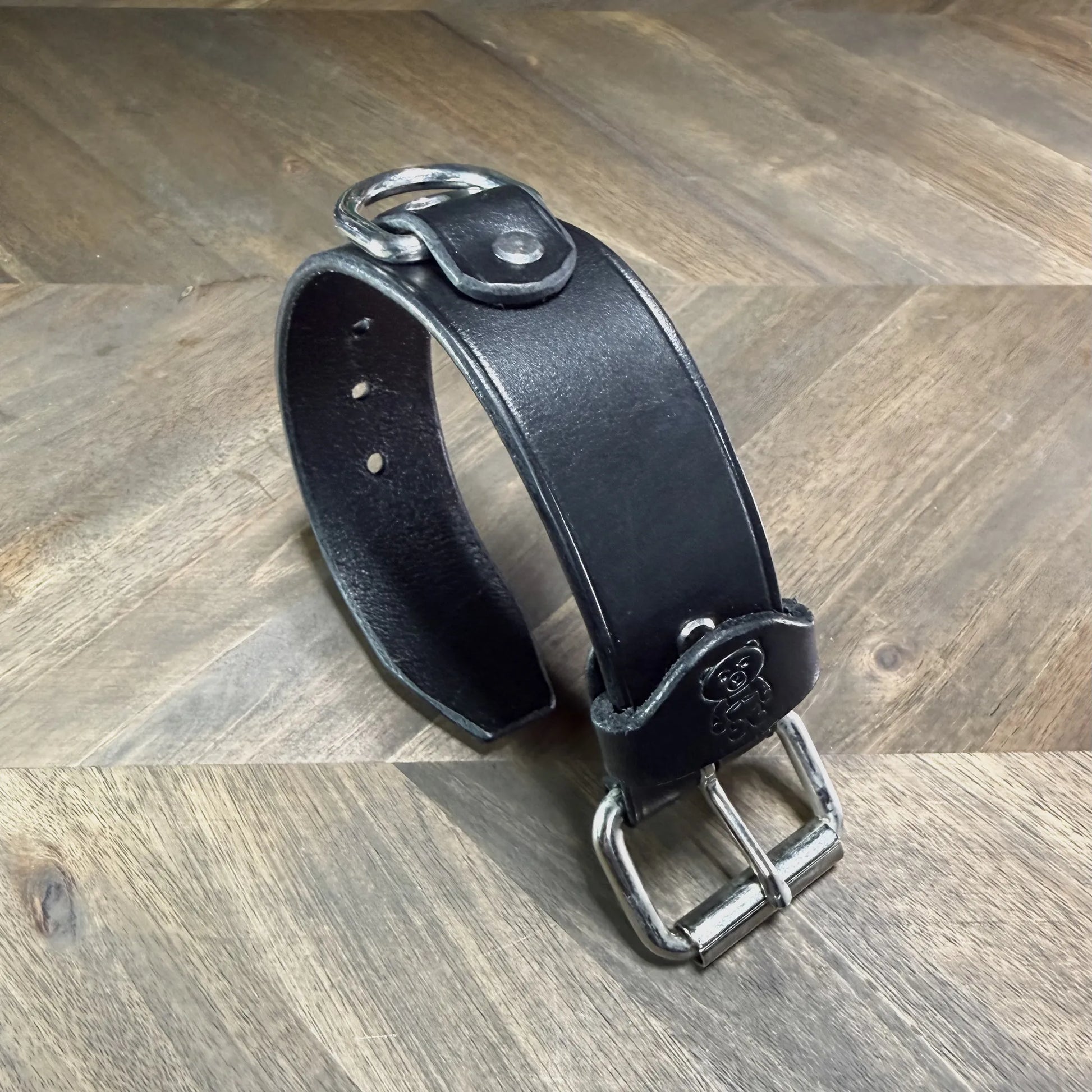 Rear perspective view of a buckled 1.5" black leather cuff highlighting a heavy-duty nickel-plated steel D-ring secured by a leather overlay.