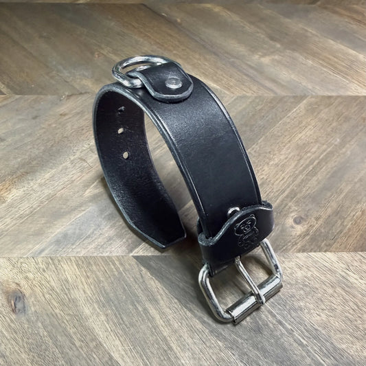 Rear perspective view of a buckled 1.5" black leather cuff highlighting a heavy-duty nickel-plated steel D-ring secured by a leather overlay.