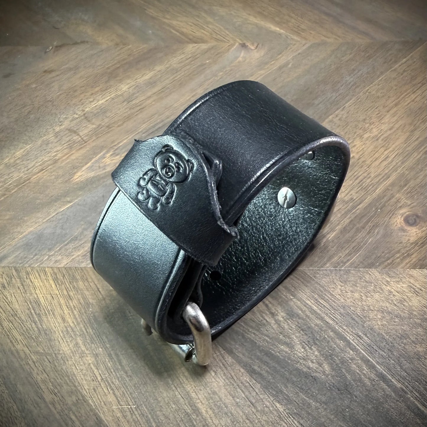 Three-quarter hero view of a buckled 1.5" black leather cuff, highlighting a debossed cartoon bear emblem on the strap end and steel rivets.