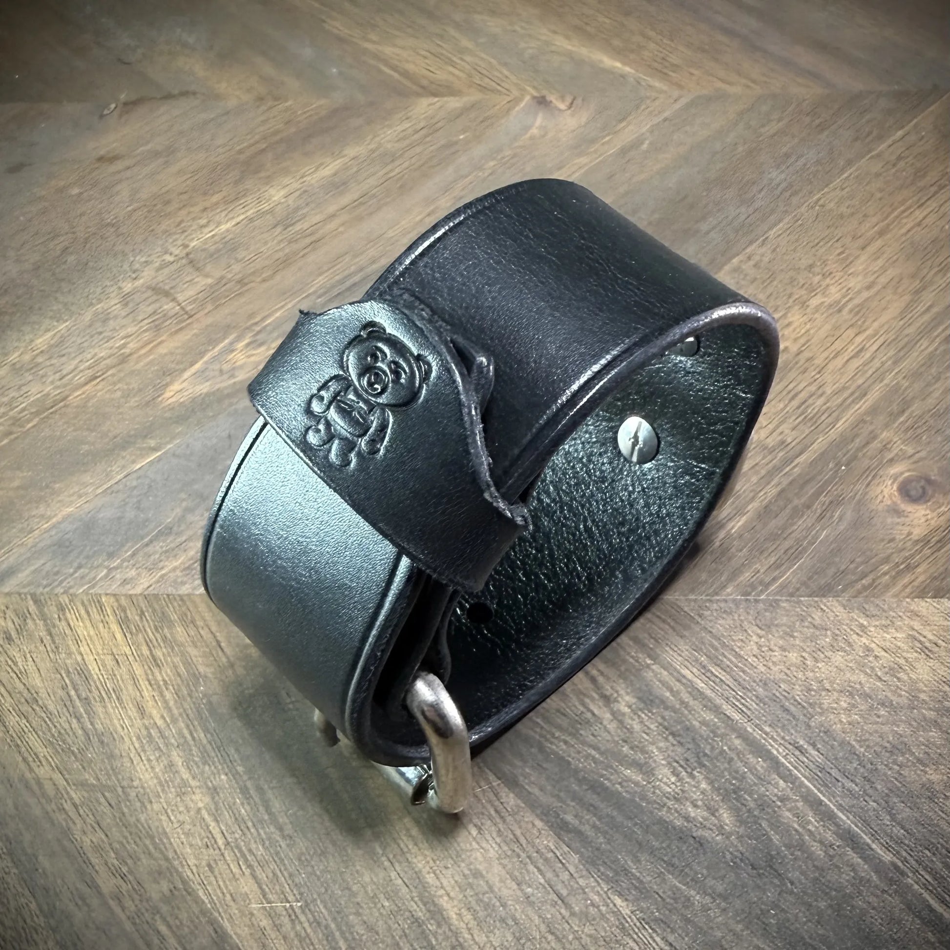 Three-quarter hero view of a buckled 1.5" black leather cuff, highlighting a debossed cartoon bear emblem on the strap end and steel rivets.