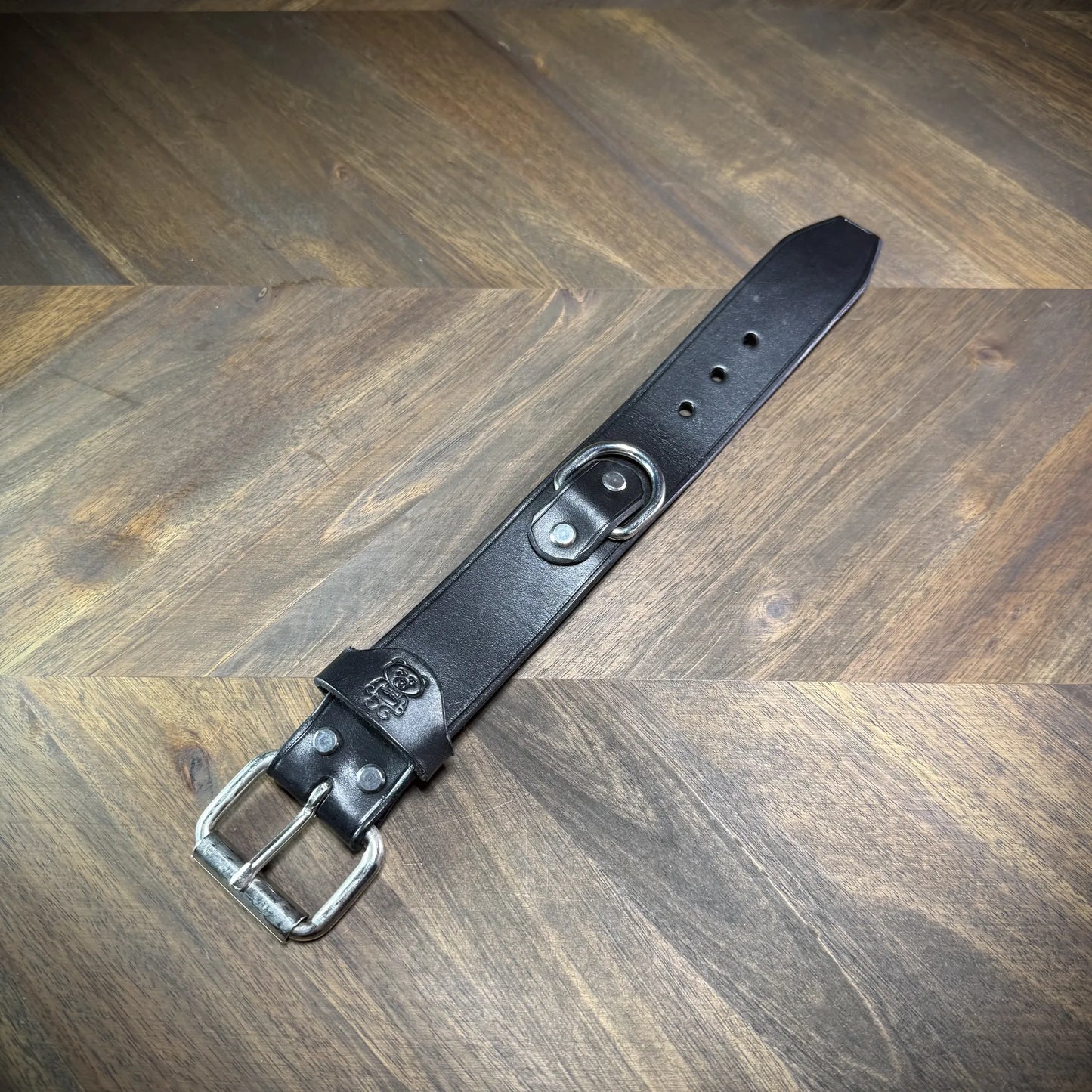 Overhead flat lay view of an unbuckled 1.5" black leather cuff with a silver-toned steel buckle, matching keeper, and centered D-ring.