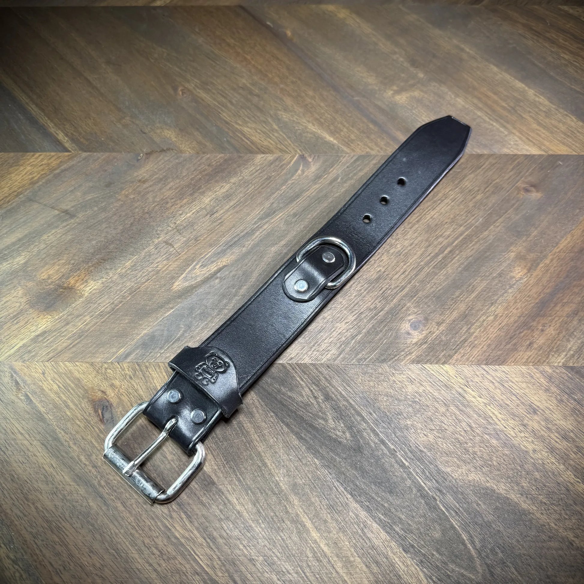 Overhead flat lay view of an unbuckled 1.5" black leather cuff with a silver-toned steel buckle, matching keeper, and centered D-ring.