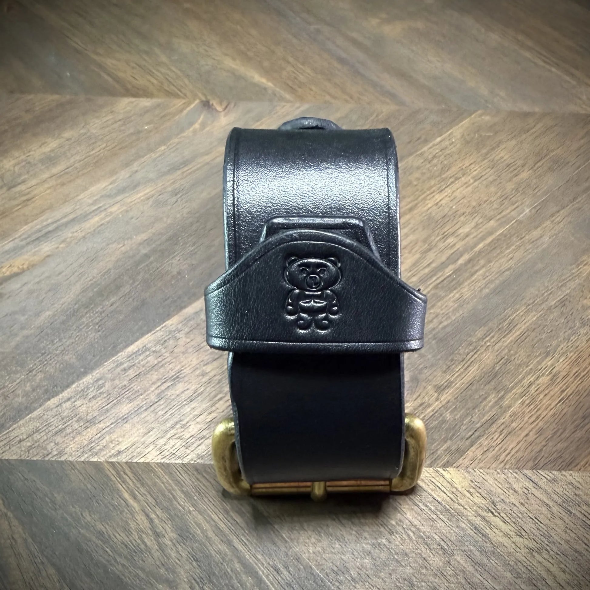 Front view of a handcrafted 1.5" leather cuff featuring thick black leather strapwork and a debossed character emblem centered above a solid brass roller buckle.