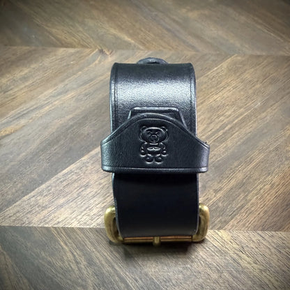 Front view of a handcrafted 1.5" leather cuff featuring thick black leather strapwork and a debossed character emblem centered above a solid brass roller buckle.