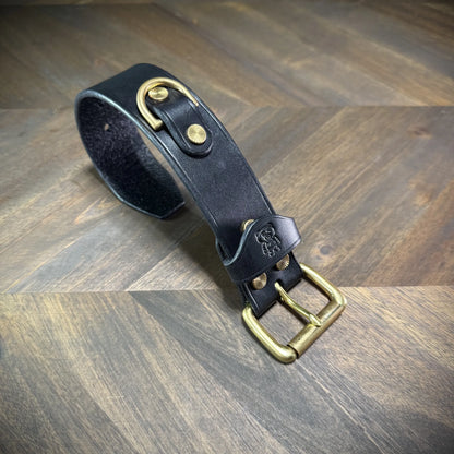 Overhead view of an unbuckled 1.5" black leather wrist cuff showcasing an integrated solid brass D-ring anchored by dual textured brass rivets and a matching roller buckle.