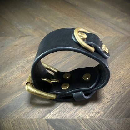 Elevated view of a closed 1.5" black leather cuff highlighting a solid brass D-ring secured with decorative brass hardware along the structural strap circumference.