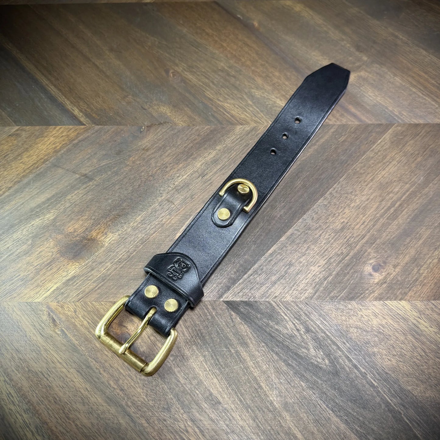 Flat lay view of a 1.5" black leather cuff extended linearly, showing the buckle attachment, debossed emblem strap keeper, integrated D-ring, and sizing adjustment holes.