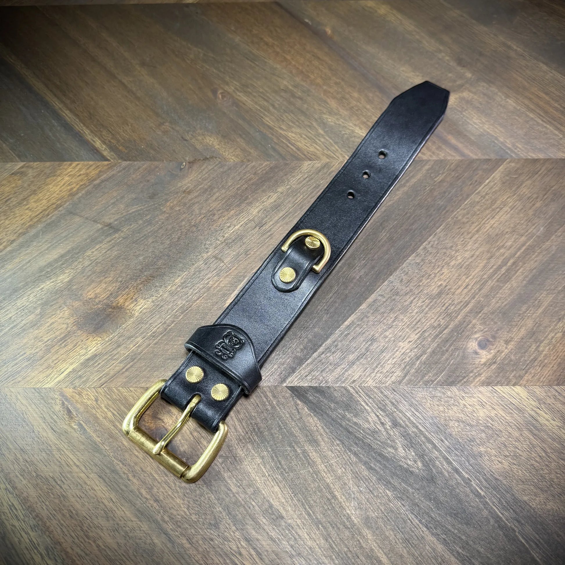 Flat lay view of a 1.5" black leather cuff extended linearly, showing the buckle attachment, debossed emblem strap keeper, integrated D-ring, and sizing adjustment holes.