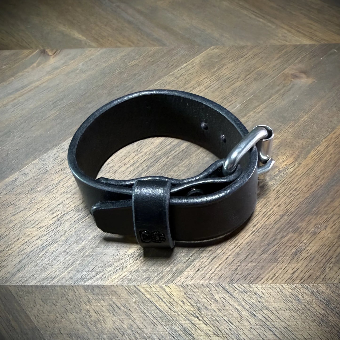 Product-reveal view of a 1.0" black leather wristband closed in a circular shape, highlighting the flush steel buckle hardware interface.