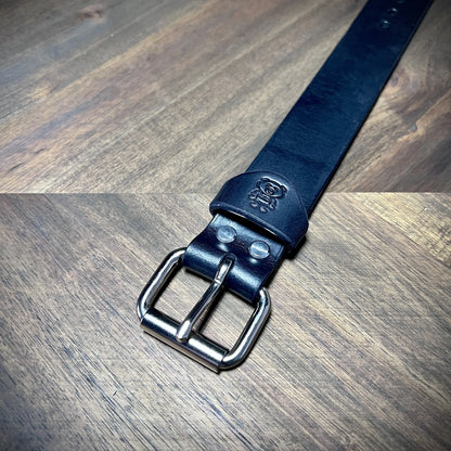 Detail macro view of a handcrafted 1.25" black leather wrist cuff highlighting a steel roller buckle and an embossed logo keeper.