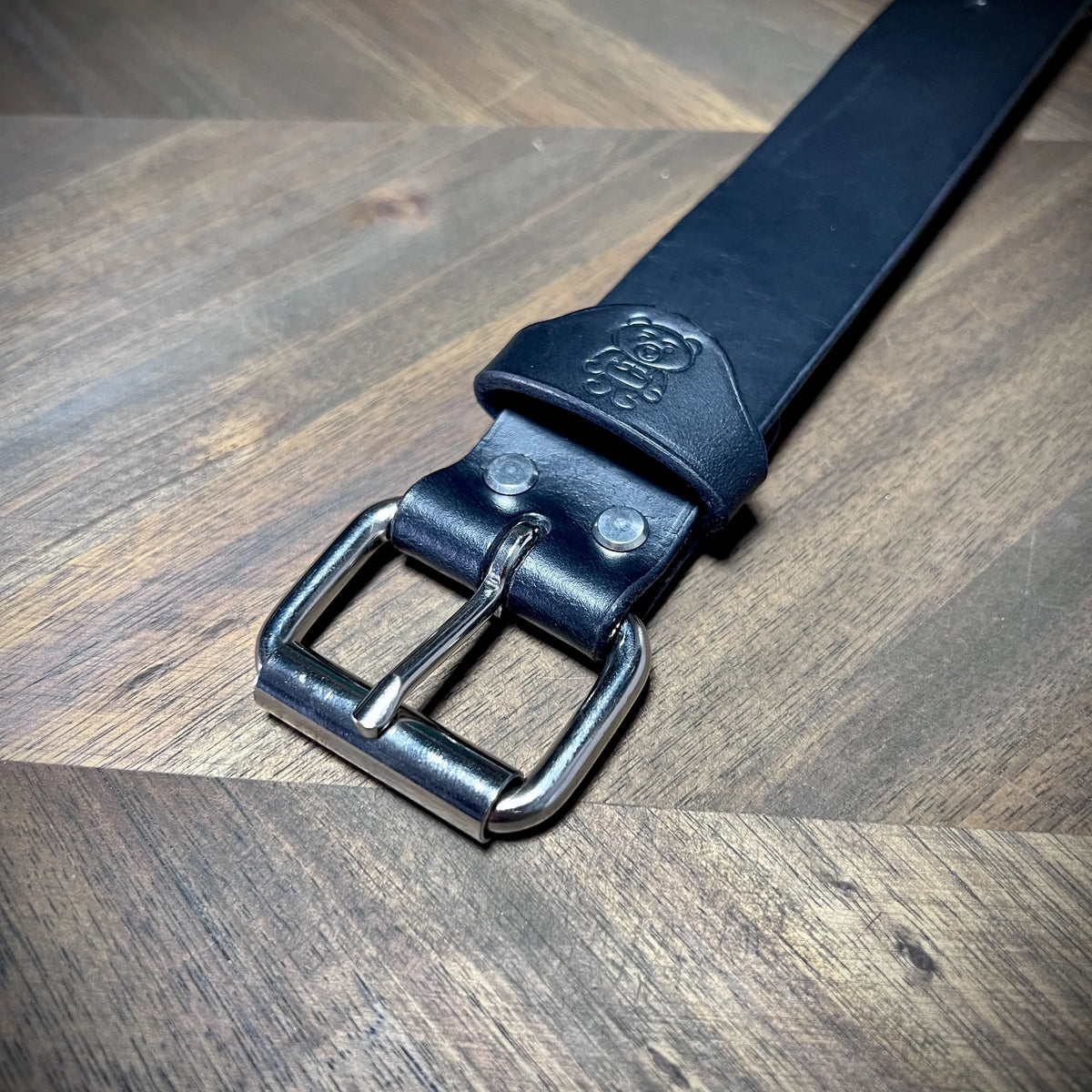 Detail macro view of the debossed bear logo and reinforced dual-rivet construction near the steel buckle of a 1.5" black leather cuff.