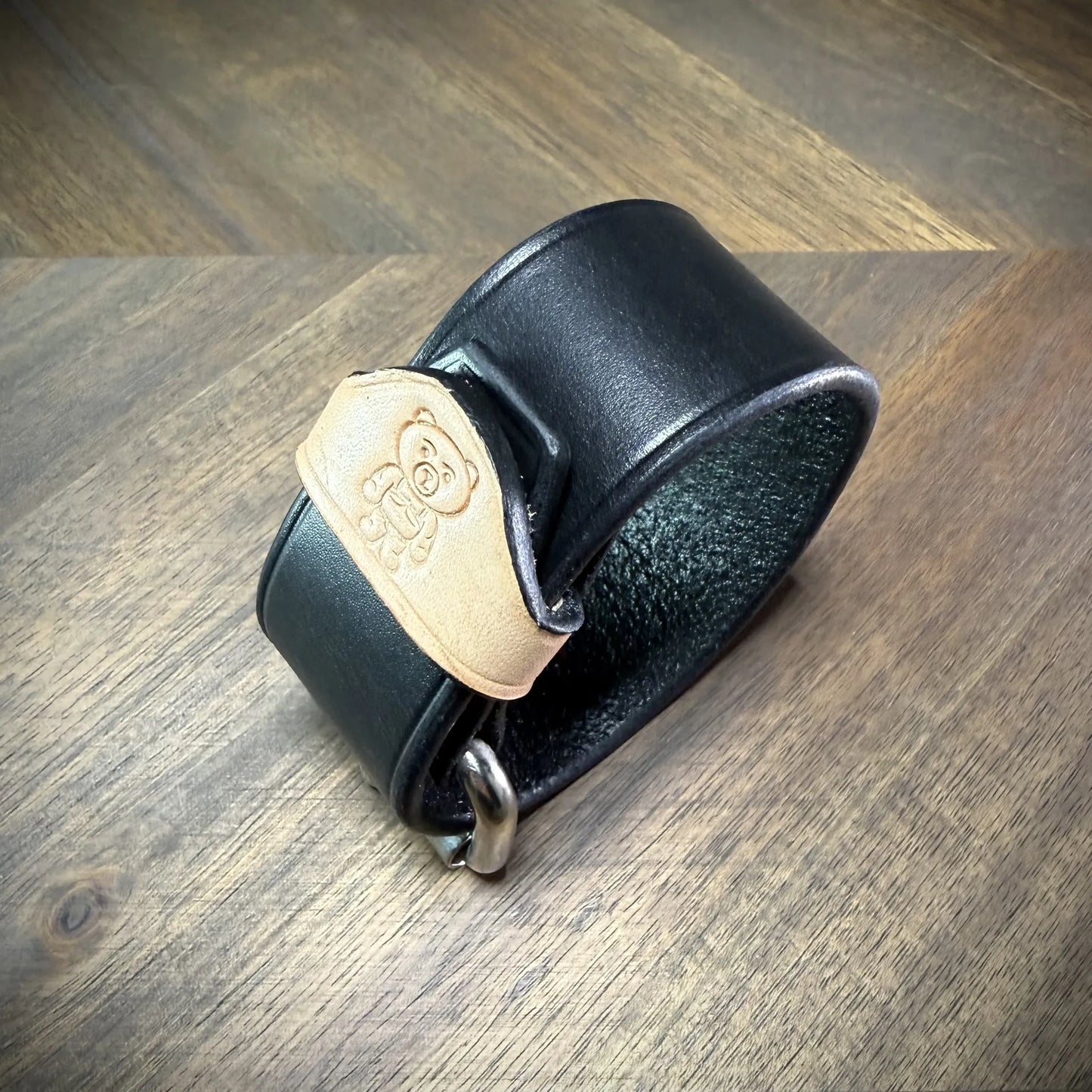 Macro detail view of a handcrafted 1.5" leather wrist cuff highlighting a natural leather belt keeper stamped with a custom bear logo.