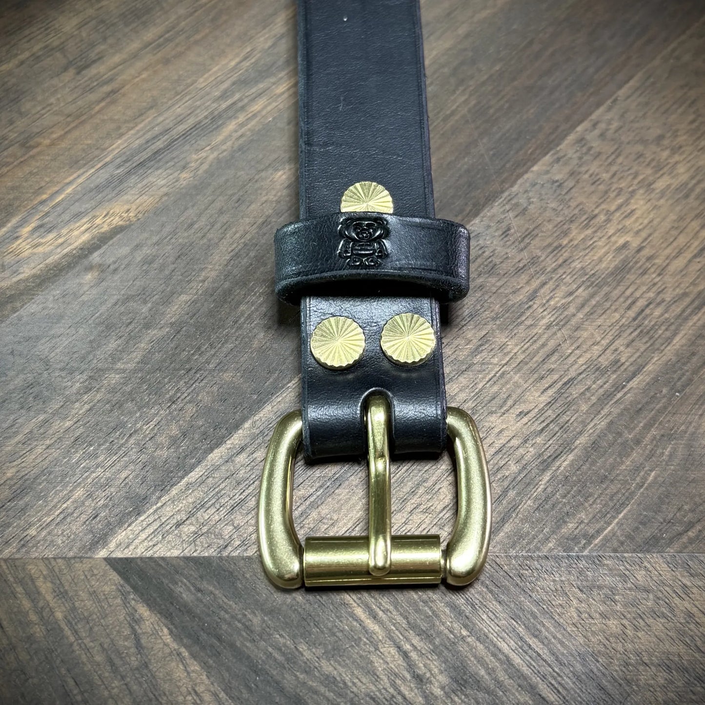 Front view of a 1" black leather cuff highlighting the vertical stack of the brass buckle, logo loop, and sunburst rivets.