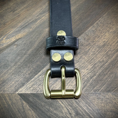 Front view of a 1" black leather cuff highlighting the vertical stack of the brass buckle, logo loop, and sunburst rivets.