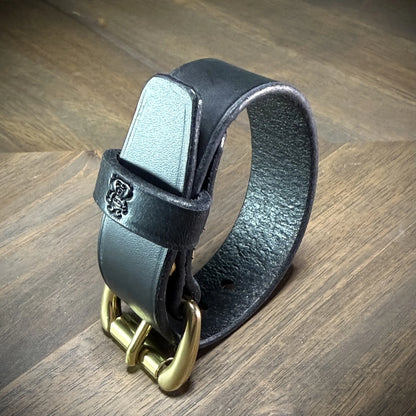 Studio profile view of a buckled 1" black leather wrist cuff standing upright to display vertical edge thickness and hardware alignment.
