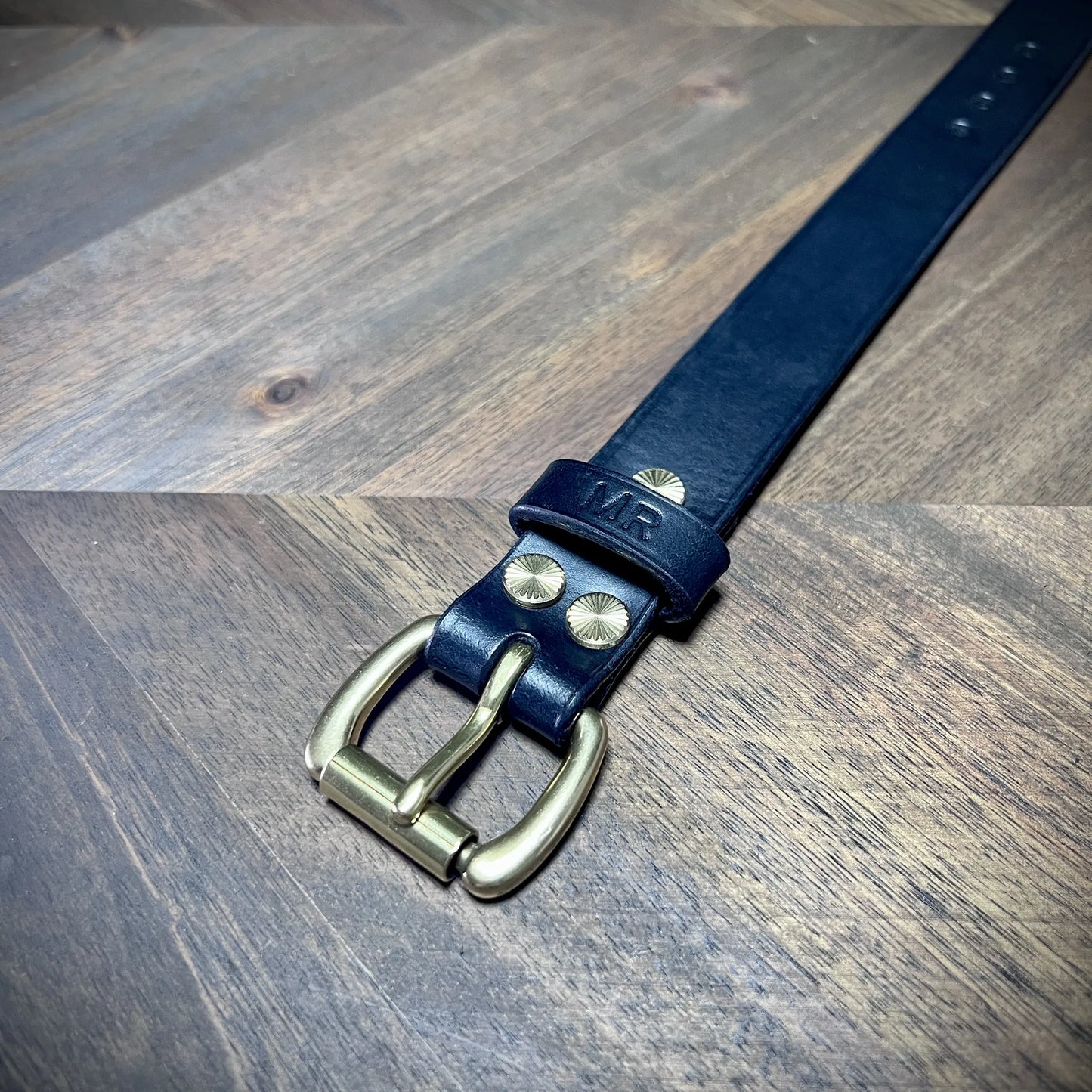 Detail macro view of a handcrafted black leather strap focusing on the solid brass buckle assembly and a personalized MR stamped keeper loop.