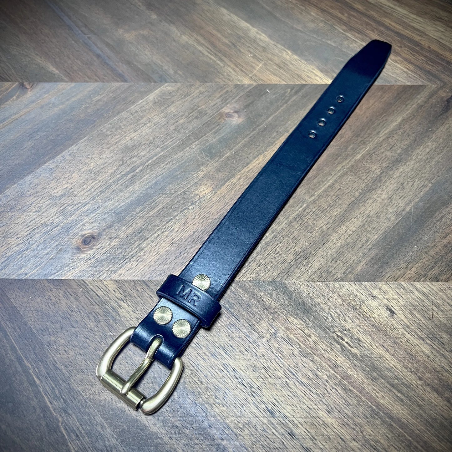 Hero tilt view of a handcrafted black leather strap showcasing a solid brass buckle, a personalized MR stamped keeper, and hand-finished edges.