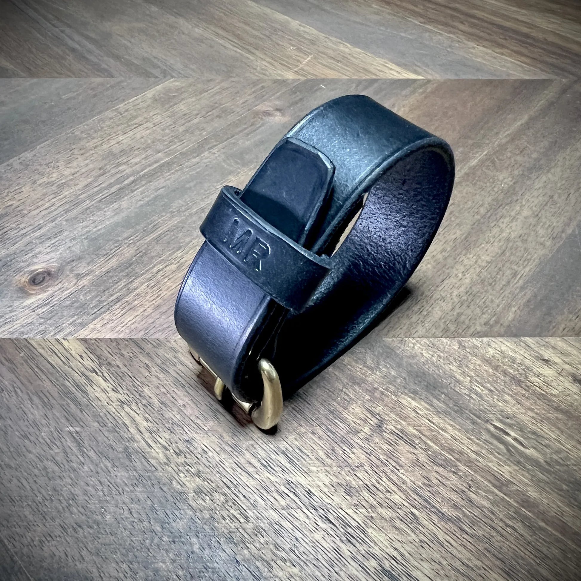 Product reveal view of a handcrafted black leather strap buckled into a closed loop, emphasizing the personalized MR stamped leather keeper.