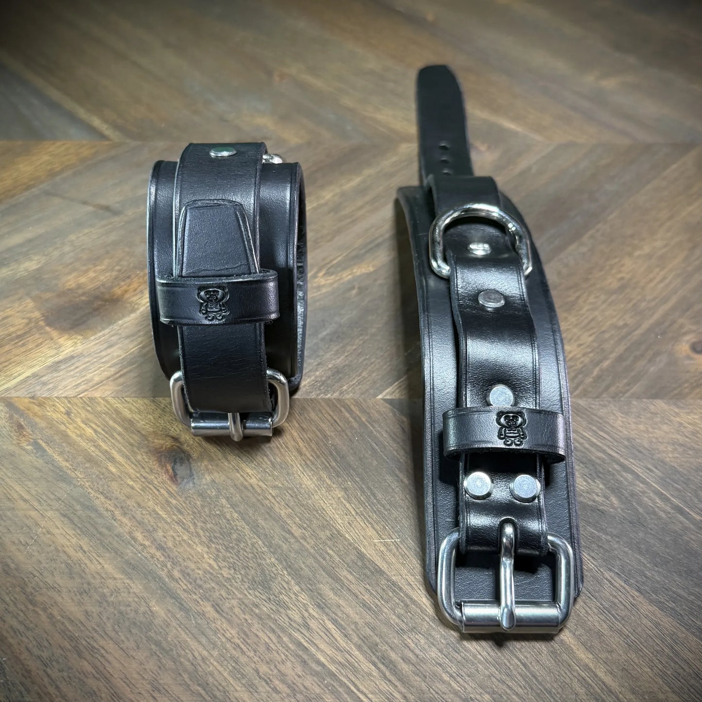 Front-facing view of two-piece black leather wrist bands with nickel-plated steel hardware, showcasing a stamped mascot emblem on the keeper.