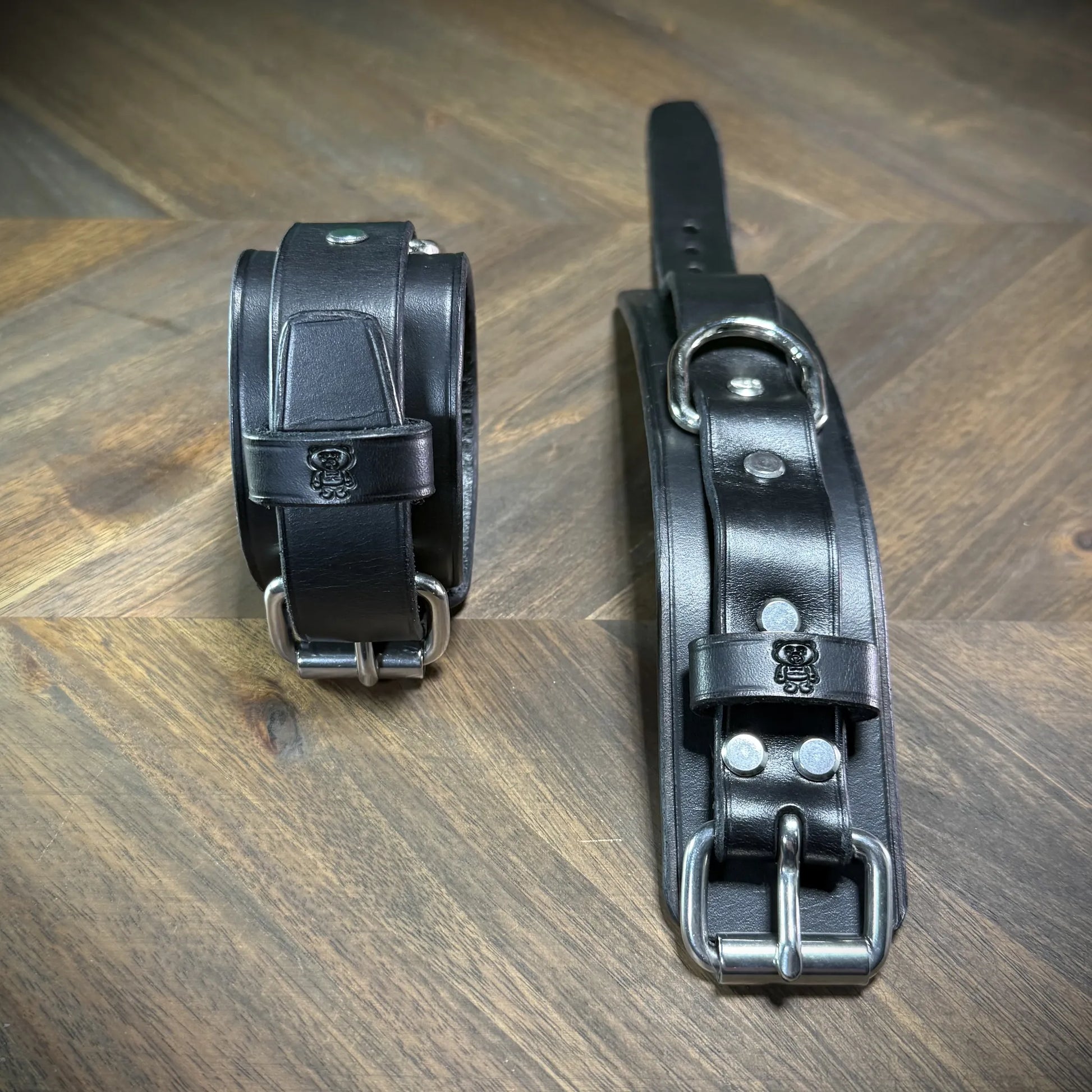 Front-facing view of two-piece black leather wrist bands with nickel-plated steel hardware, showcasing a stamped mascot emblem on the keeper.