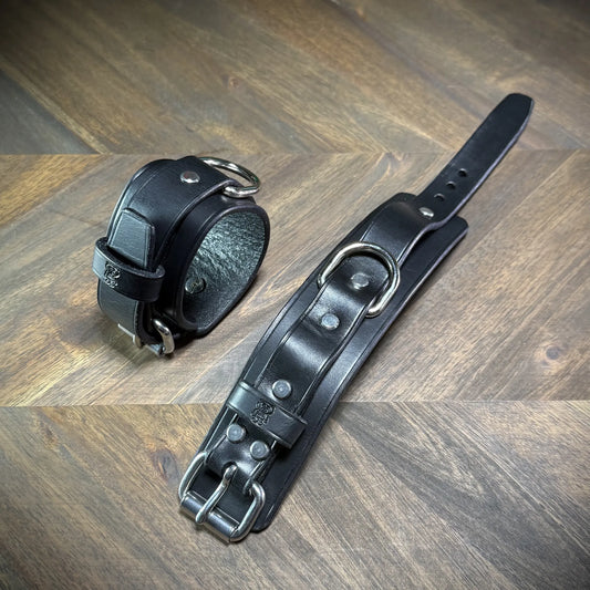 Hero-tilt view of two handcrafted 1.75" black leather two-piece cuffs with nickel-plated steel hardware buckles and centered structural D-rings.
