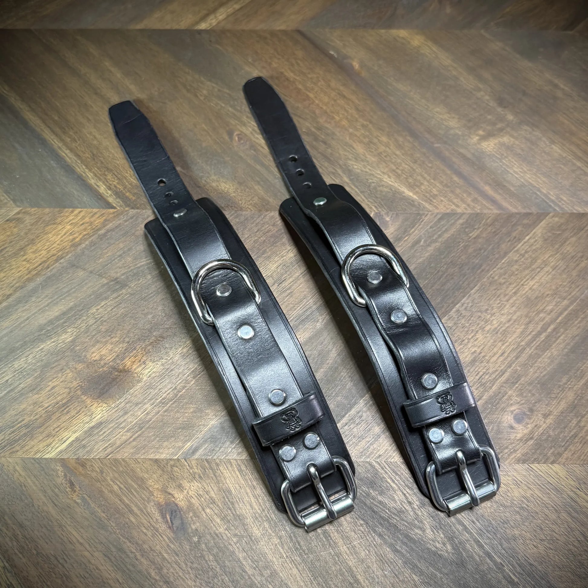 Parallel layout of two flat 1.75" black leather two-piece wrist straps revealing nickel-plated D-rings and double structural steel rivets.