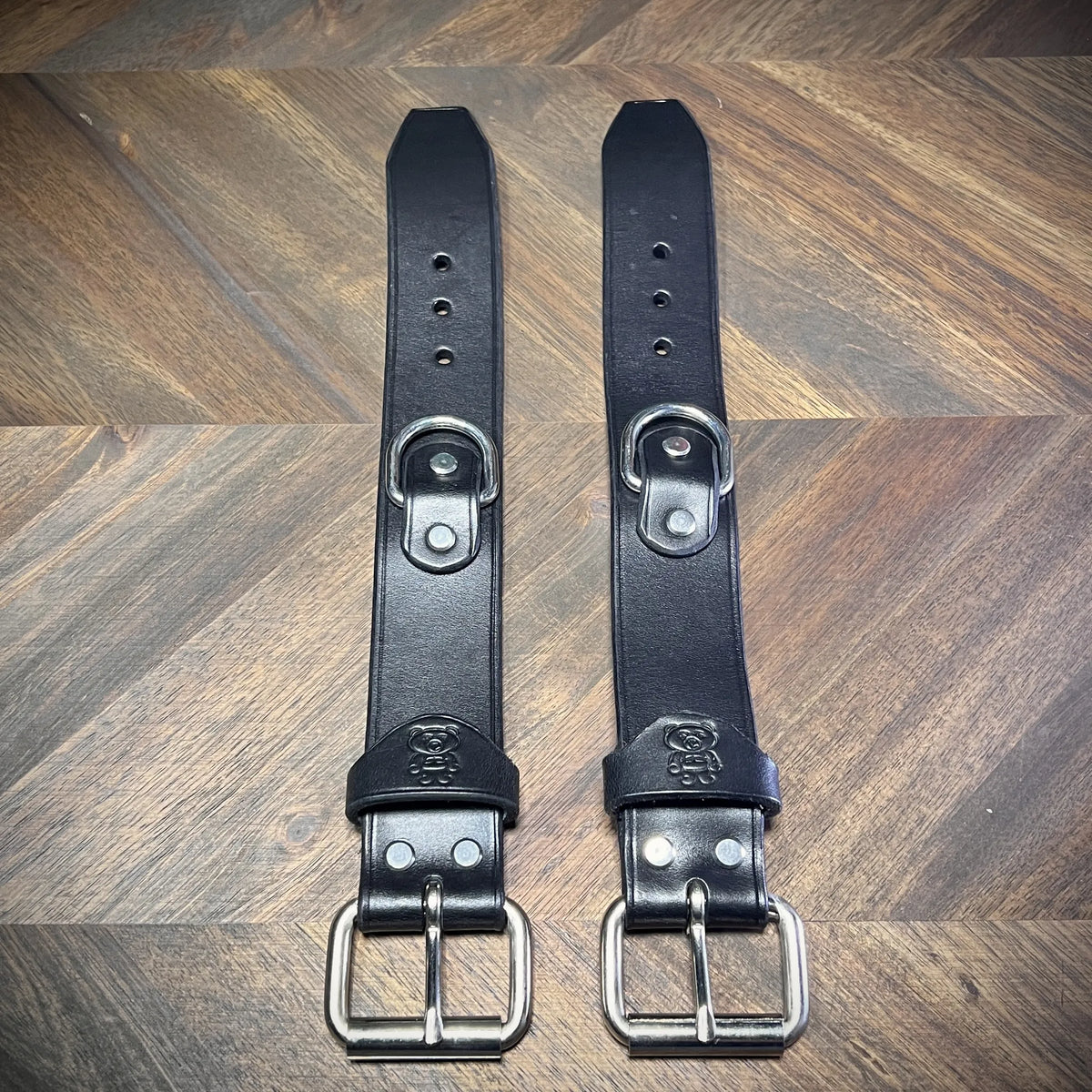 Top-down flat lay of a pair of unbuckled 1.5" black leather straps showcasing symmetric adjustment holes, centered steel D-rings, and roller buckles.